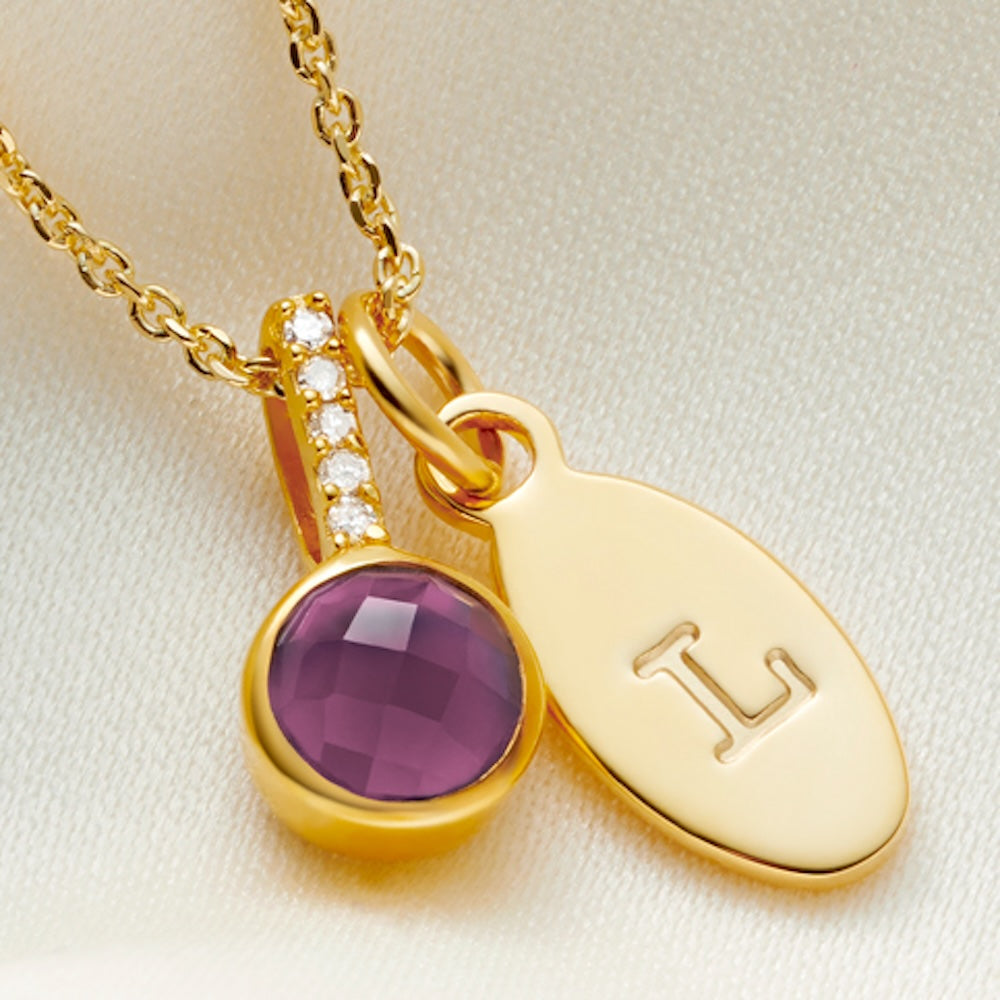 Gold necklace with a pendant charm with a cushion cut faceted purple amethyst stone in a rub over setting. There are 5 tiny white diamonds in the bale of the pendant and it's hung on a dainty gold vermeil chain along side an oval charm, hand stamped with the initial L.