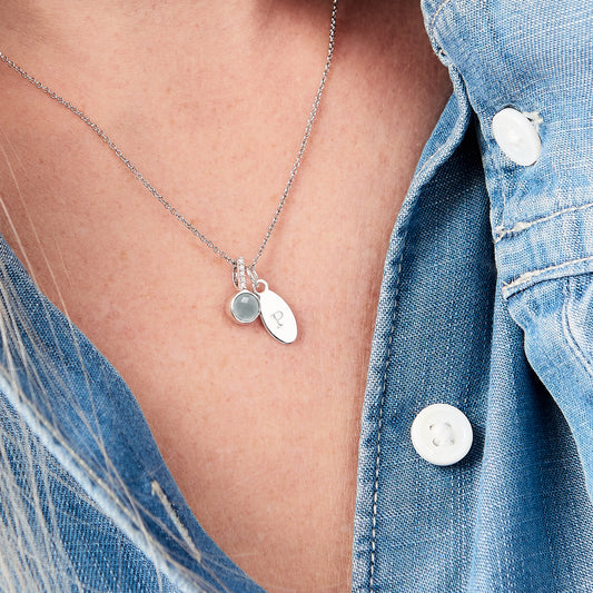 Faceted round aquamarine pendant, set in sterling silver with tiny white diamonds in the bale.  Hung alongside a silver oval charm, personalised with a capital letter P.  Both come hung on a fine trace chain by Claudette Worters.