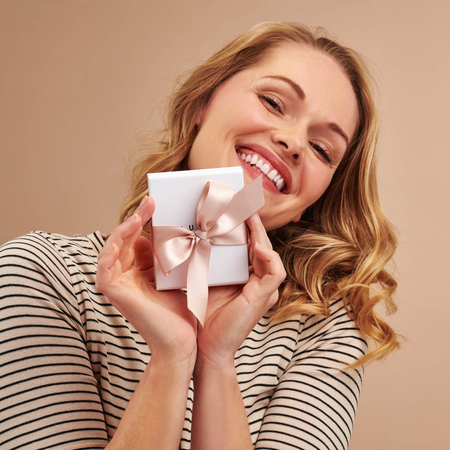 Smiling blonde woman in a striped tee holding Claudette Worters white gift box tied with a blush pink satin ribbon.