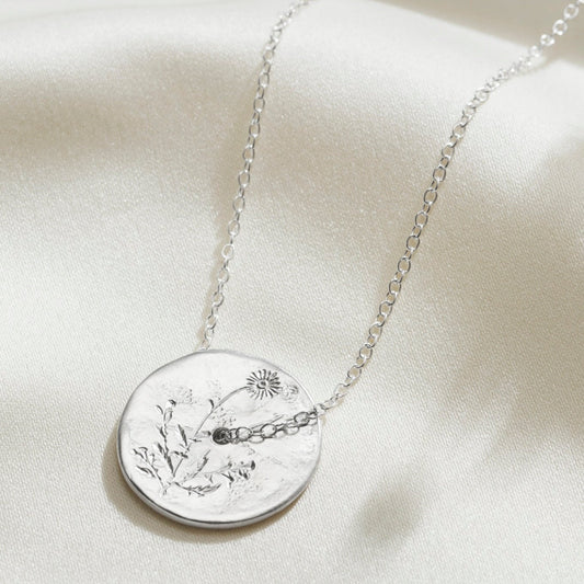 Hand engraved daisy birth flower on a sterling silver disc pendant with a hole in the middle and a fine trace chain slotting through the middle.