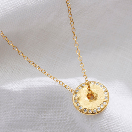 Gold necklace with a circular pendant with a ring of white diamonds symbolising a New Moon with a trace chain running through the centre on a white fabric background.