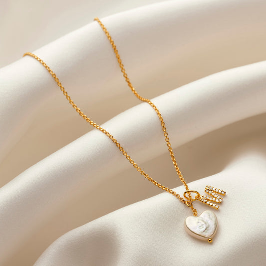 Gold necklace with a captial letter M charm that's pavé set with white diamonds.  Hung on a fine gold trace chain along side a heart shaped freshwater pearl
