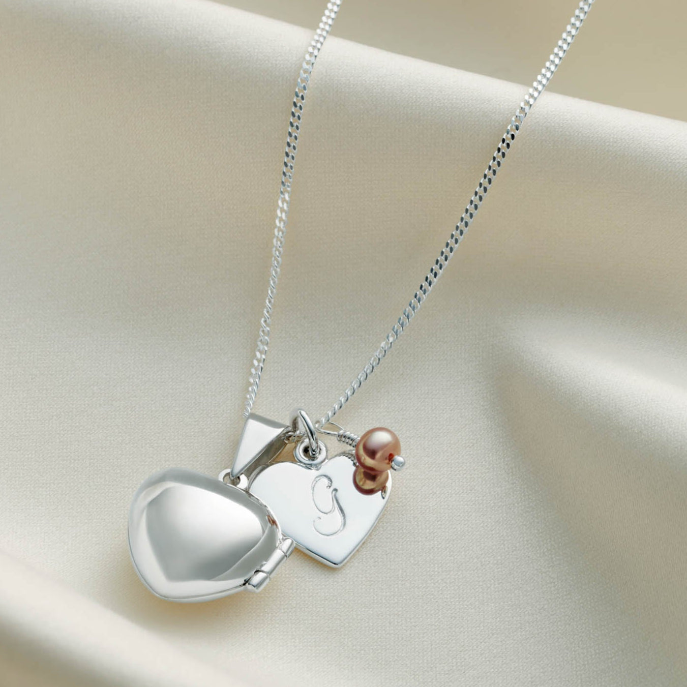 Sterling silver heart shaped locket necklace with a heart charm with the letter G hand stamped on it.  There's also a pink freshwater pearl birthstone charm on the necklace and al the elements are hung on a sterling silver chain.