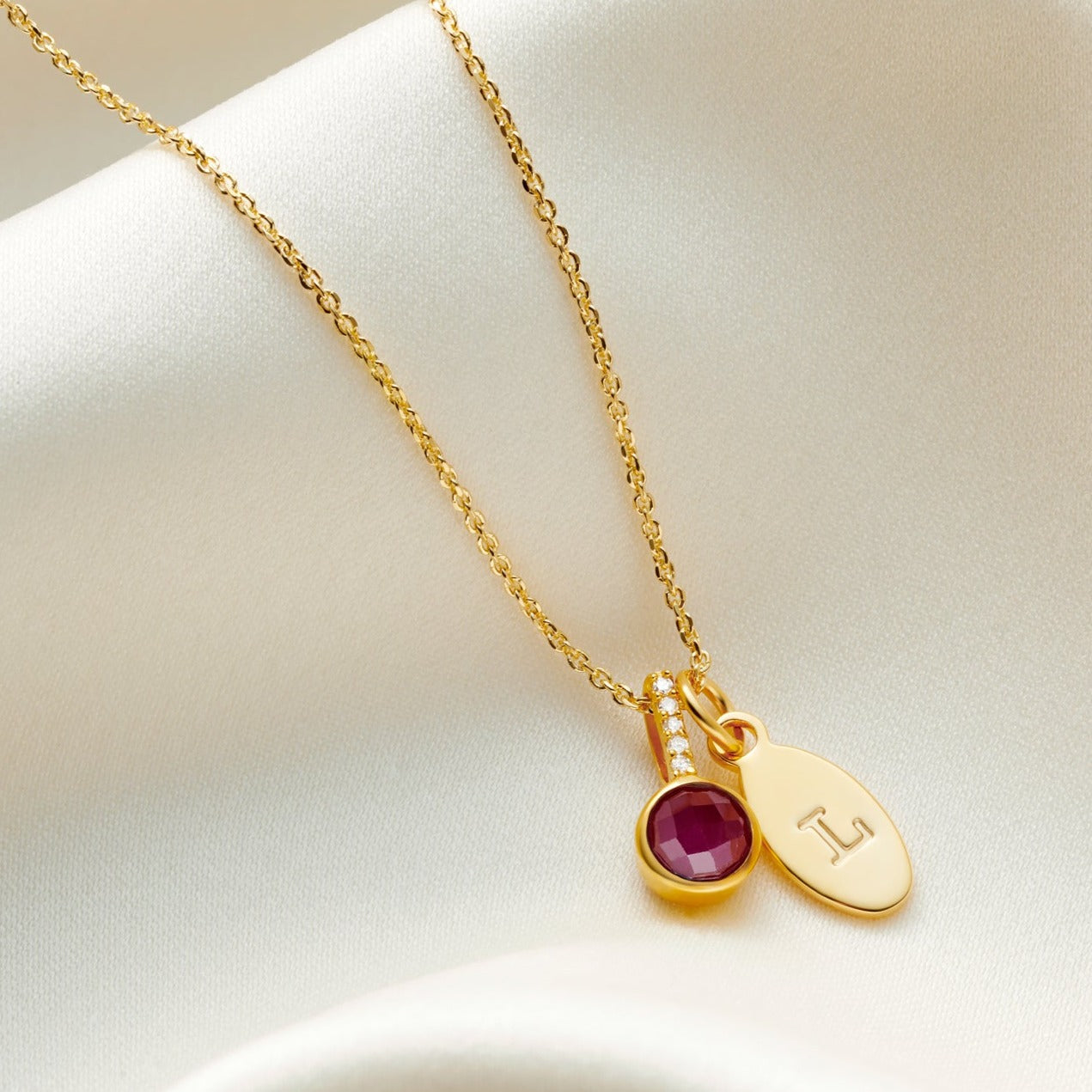 Faceted ruby gemstone pendant in 18 carat yellow gold with tiny diamonds in the bale on a fine trace chain with a small oval charm personalised with the letter L.