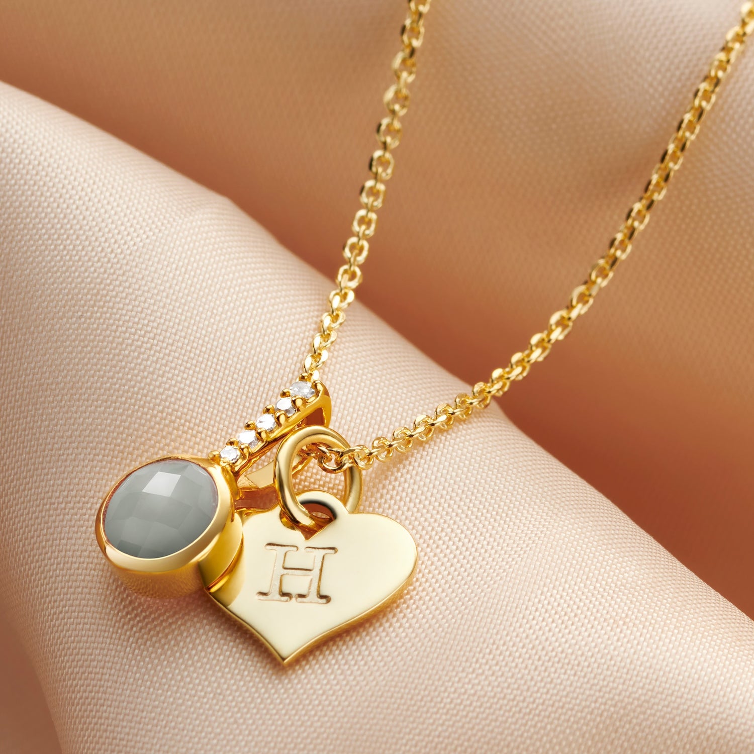 Gold vermeil pendant with a single round faceted aquamarine stone in a rub over setting with tiny white diamonds in the bale.  The pendant is on a fine gold vermeil trace chain, next to a gold heart charm hand stamped with the letter H.