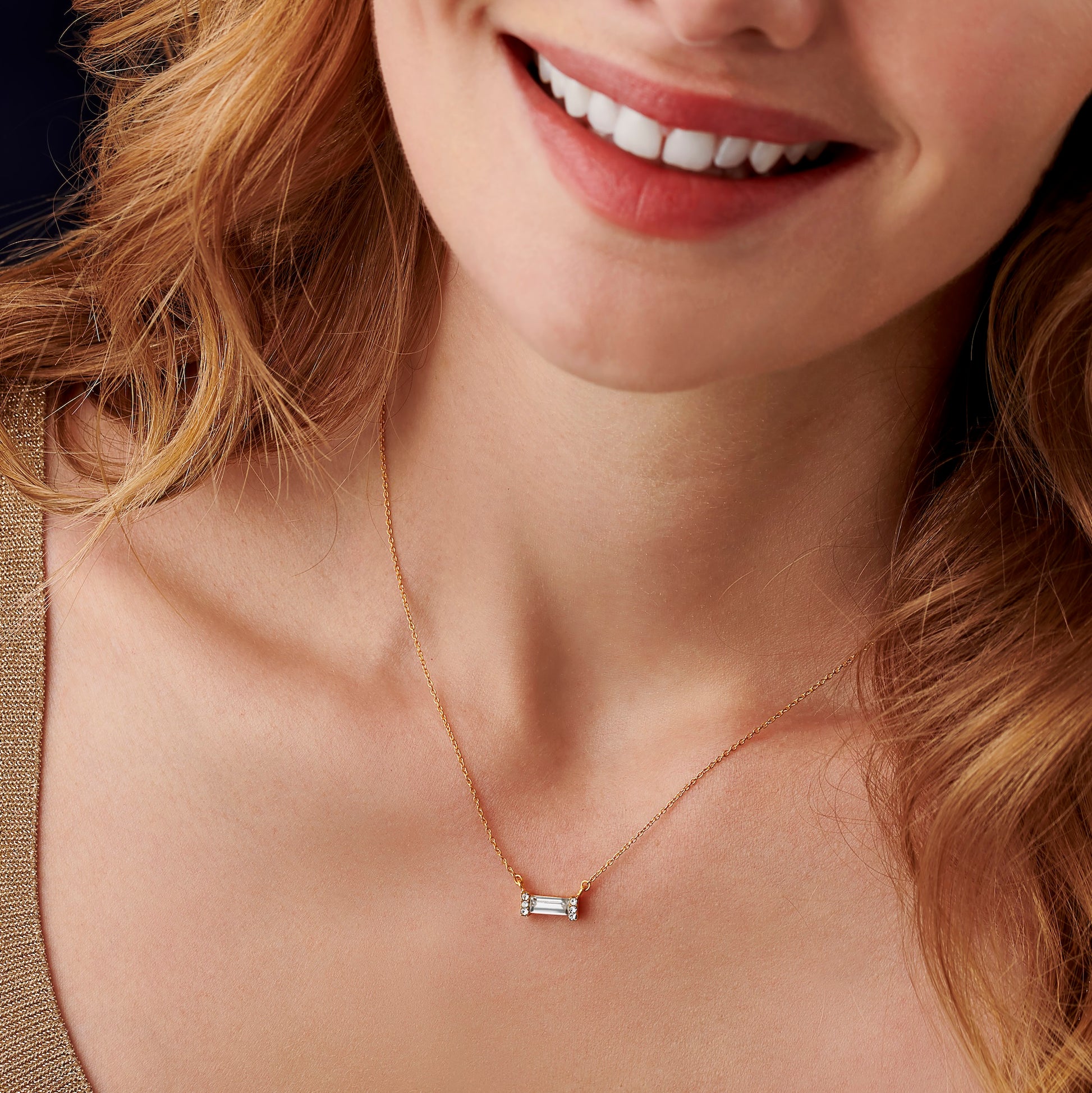 Smiling woman wearing a dainty gold necklace with a baguette cut lear crystal, surrounded by tiny white crystals suspended from a fine trace chain.