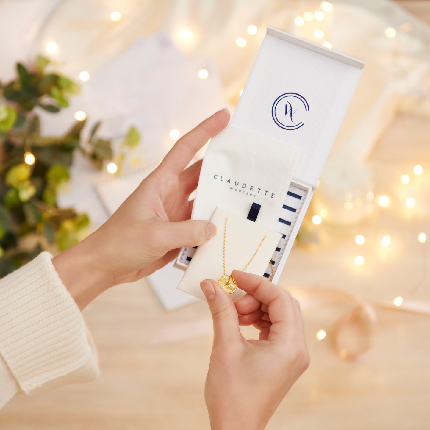 Person holding a  white gift box with 'Claudette Worters' branding against a blurred background with lights and greenery.