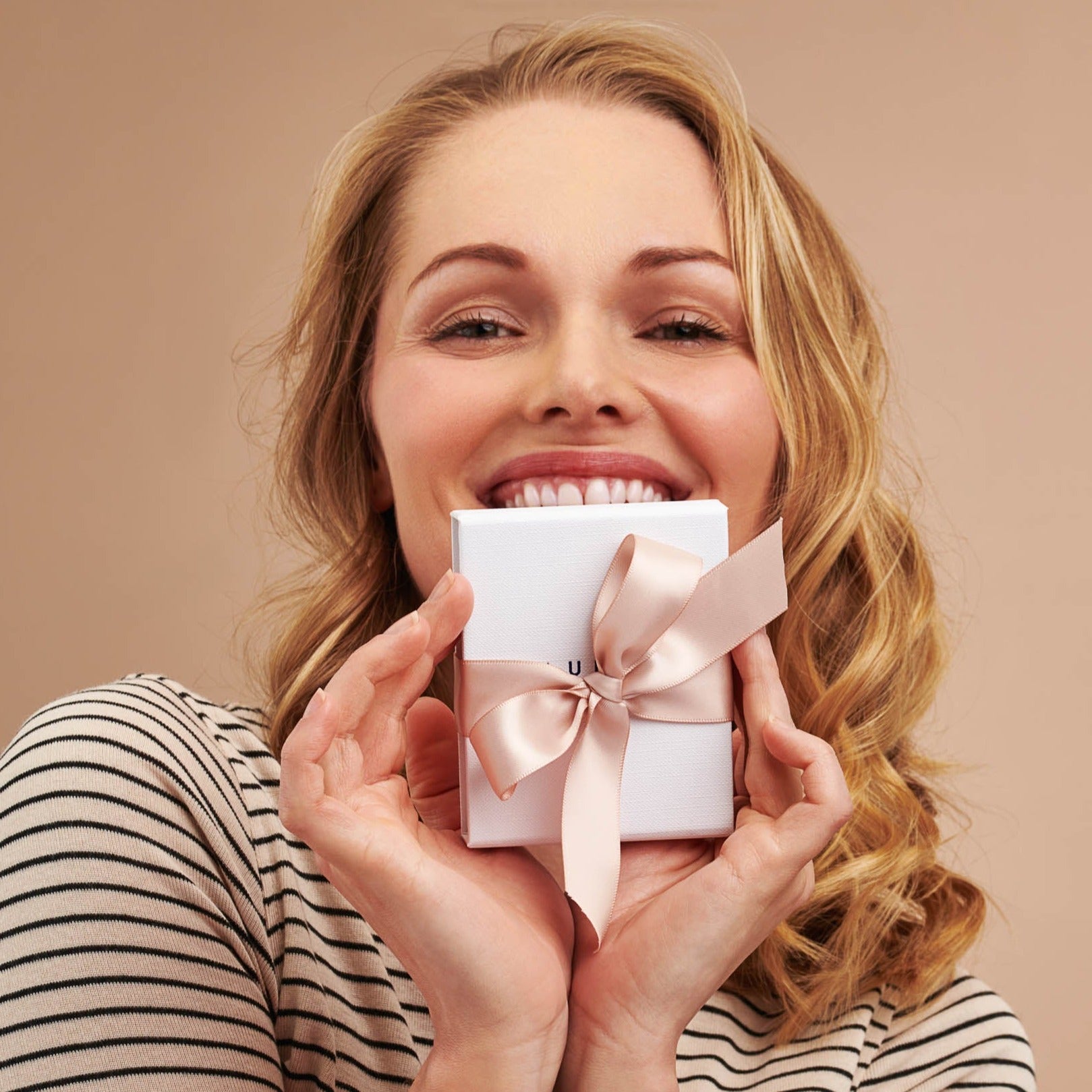 Woman in a striped top smiling, holding white gift box tied with a blush pink satin bow.