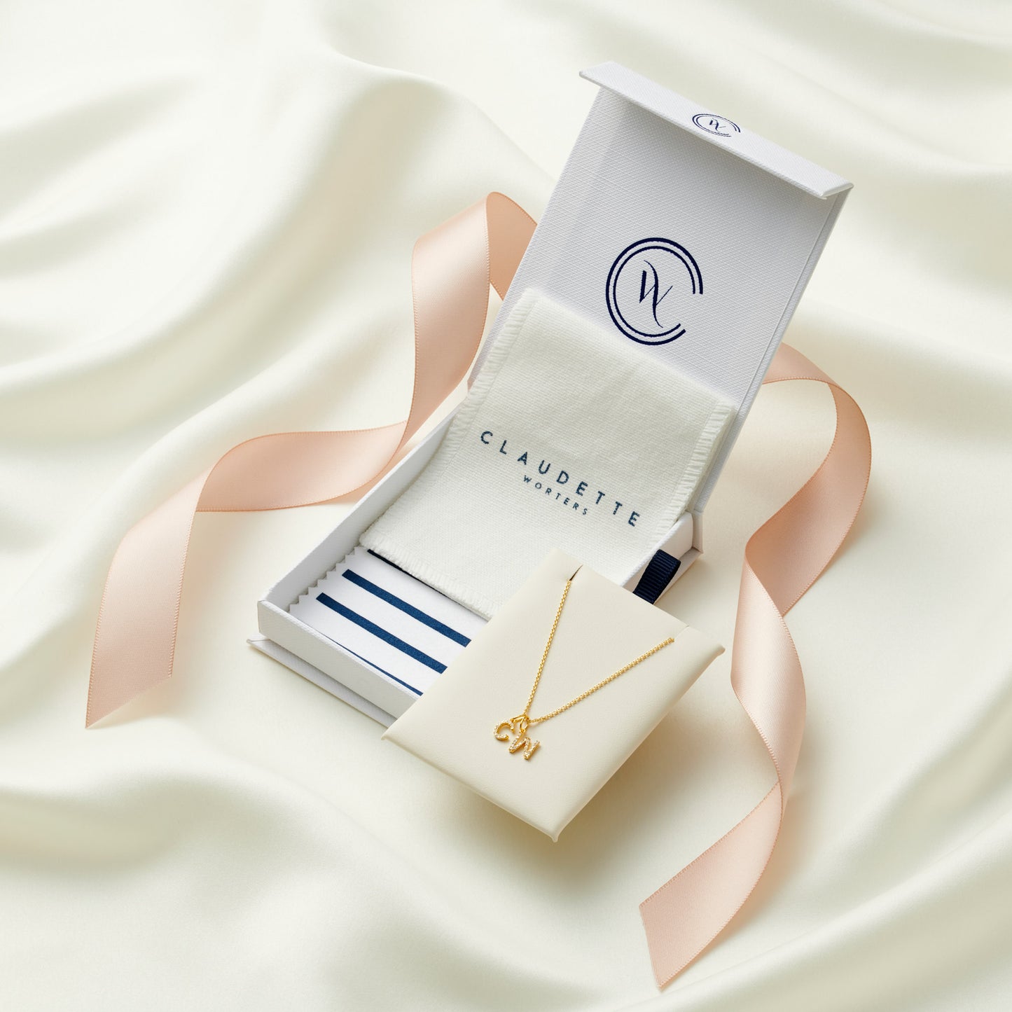 Claudette Worters jewellery gift box packaging with white gift box, blush pink satin ribbon, linen pouch and striped polishing cloth and a cream faux leather insert pad with a diamond letter pendant.