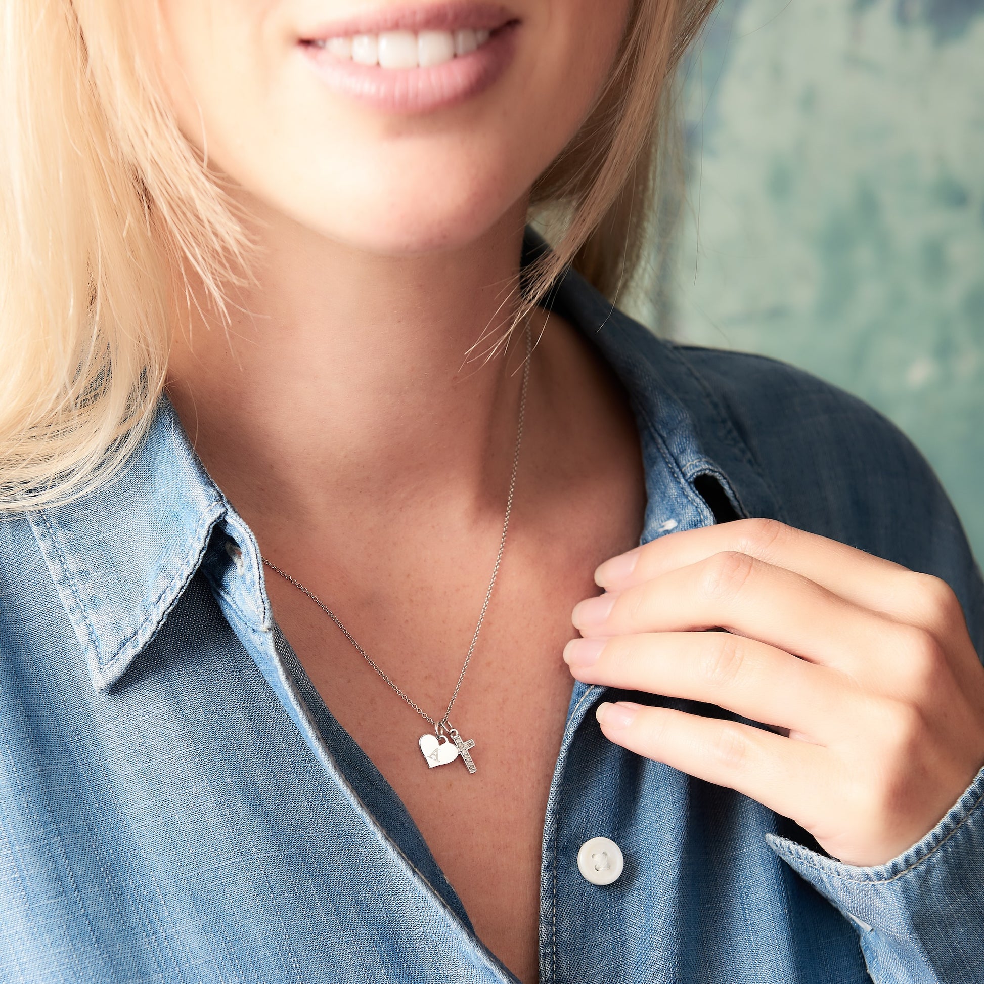 Female model wearing a cross necklace featuring a small sterling silver cross set with white diamonds along side a heart charm hand stamped with the letter A on a fine silver trace chain.