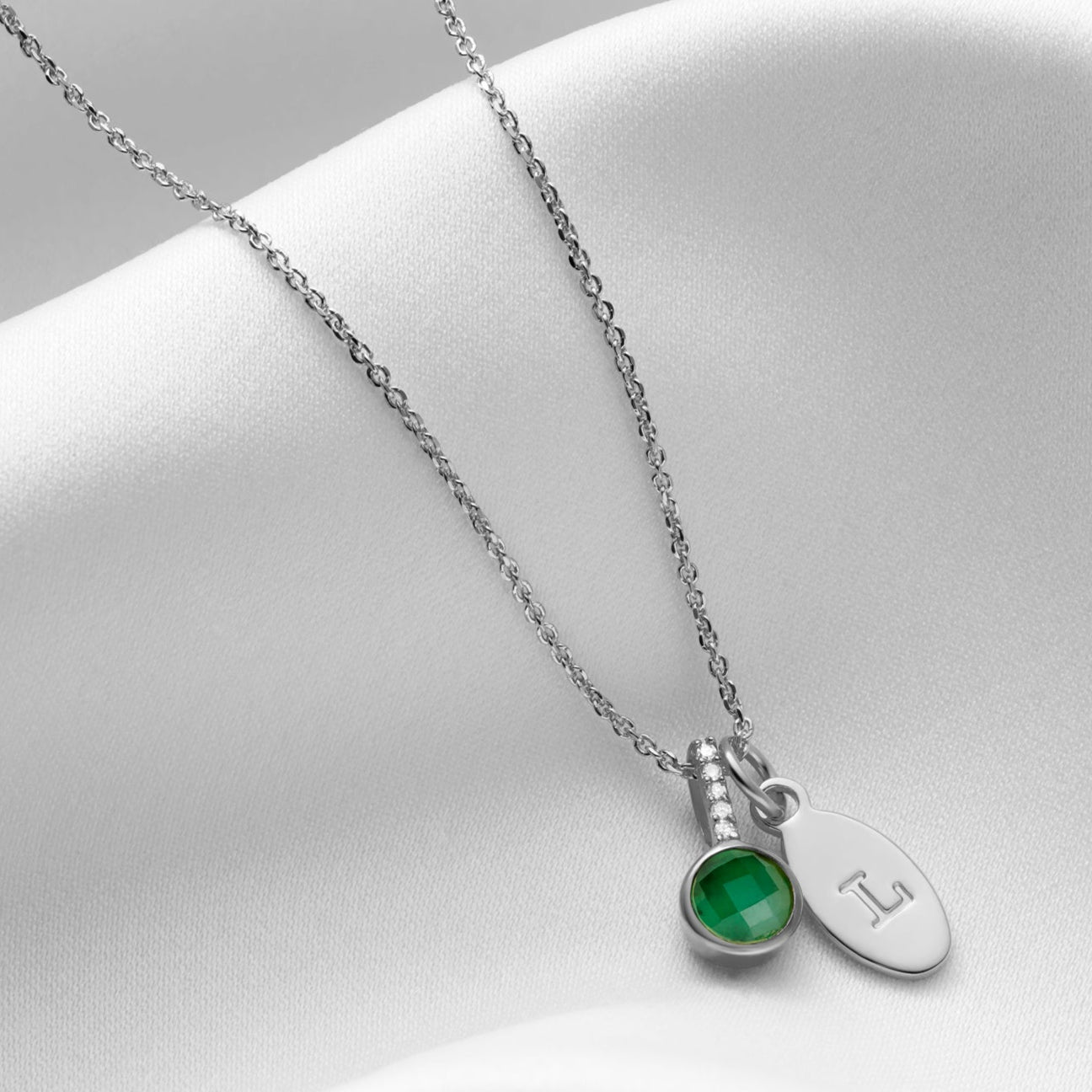 Emerald gemstone necklace in silver.  Necklace has a round, cushion cut green emerald in a sterling silver setting with tiny white diamonds in the bale.  There is a silver oval charm personalised with the letter L which is hung along side the emerald pendant on a fine sterling silver trace chain.  The piece is by Claudette Worters.