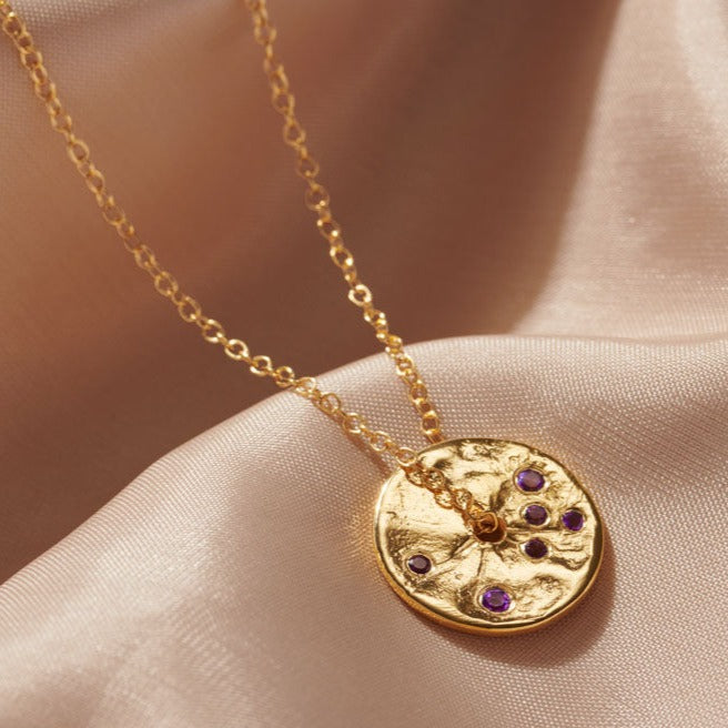 Round disc pendant in gold with a molten texture.  Pendant is randomly set with six purple amethyst stones.  There's a hole in the middle of the pendant and a fine gold trace chain is running through the middle so that the pendant can rotate.