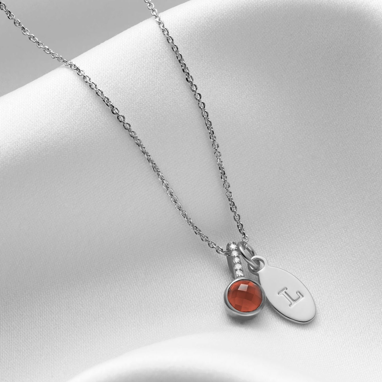 Silver pendant necklace set with a faceted round dark red garnet with tiny white diamonds in the bale.  The garnet pendant is hung along side a silver oval charm that has been hand stamped with a capital letter L.  Both hung on a delicate sterling silver trace chain.
