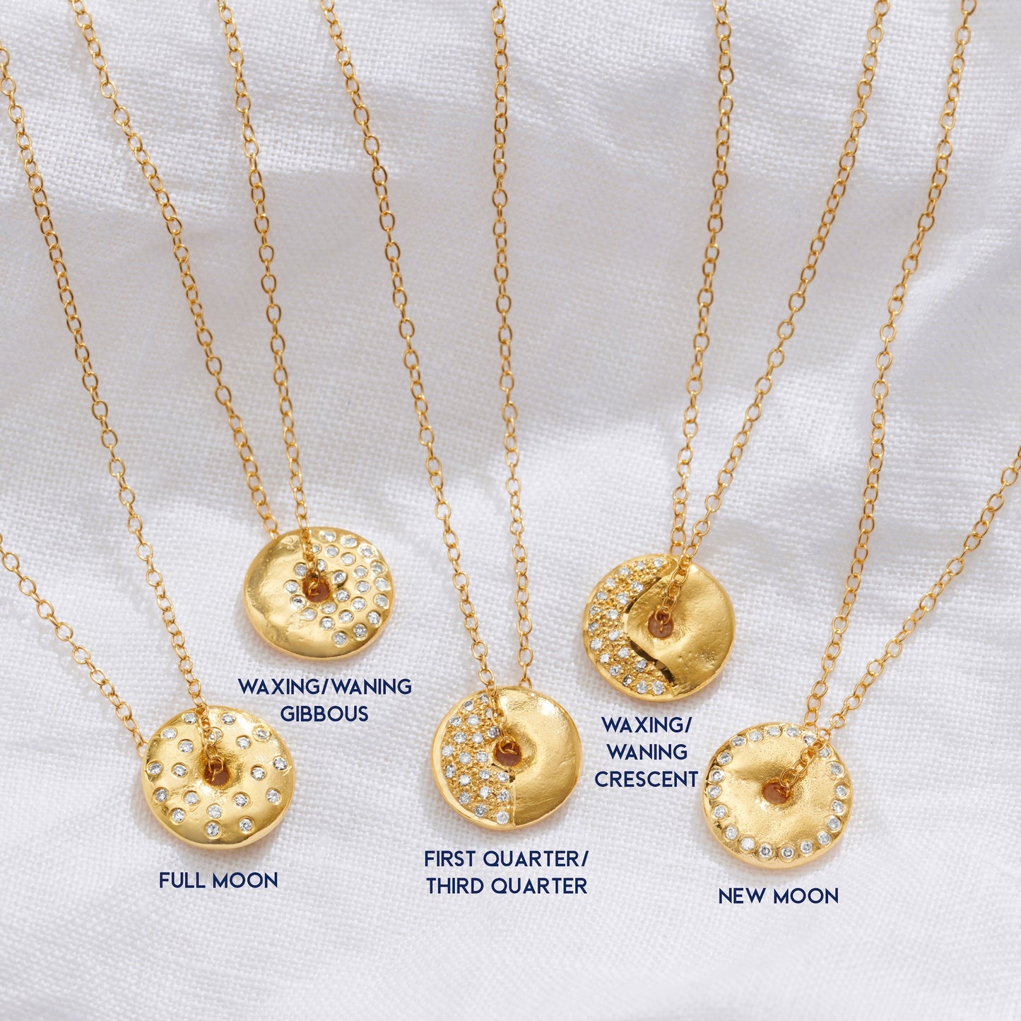 Gold necklaces with circular pendants with a textured surface, set with diamonds labeled for different phases of the moon on a white background by Claudette Worters