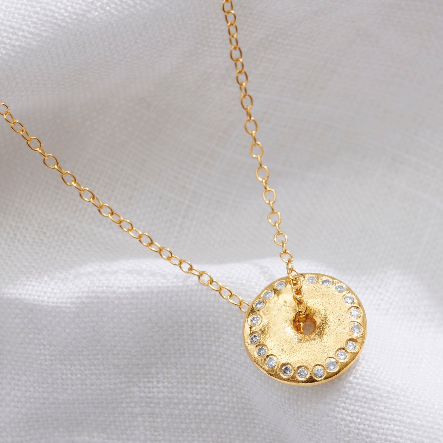 Gold necklace with a circular pendant with a ring of white diamonds symbolising a New Moon with a trace chain running through the centre on a white fabric background.