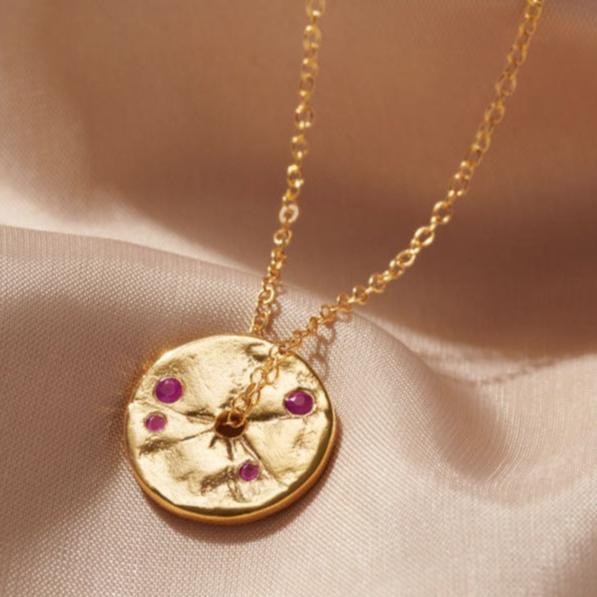 Gold disc pendant with textured surface like an ancient coin.  The pendant is set with faceted rubies.  It has a hole in the centre of the pendant and a dainty trace chain runs through the middle.