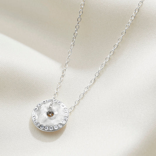 Sterling silver necklace with a circular pendant with a hole in the middle with a fine chain running through the centre on a cream background.  The edge of the pendant is set with a ring of small white diamonds by Claudette Worters