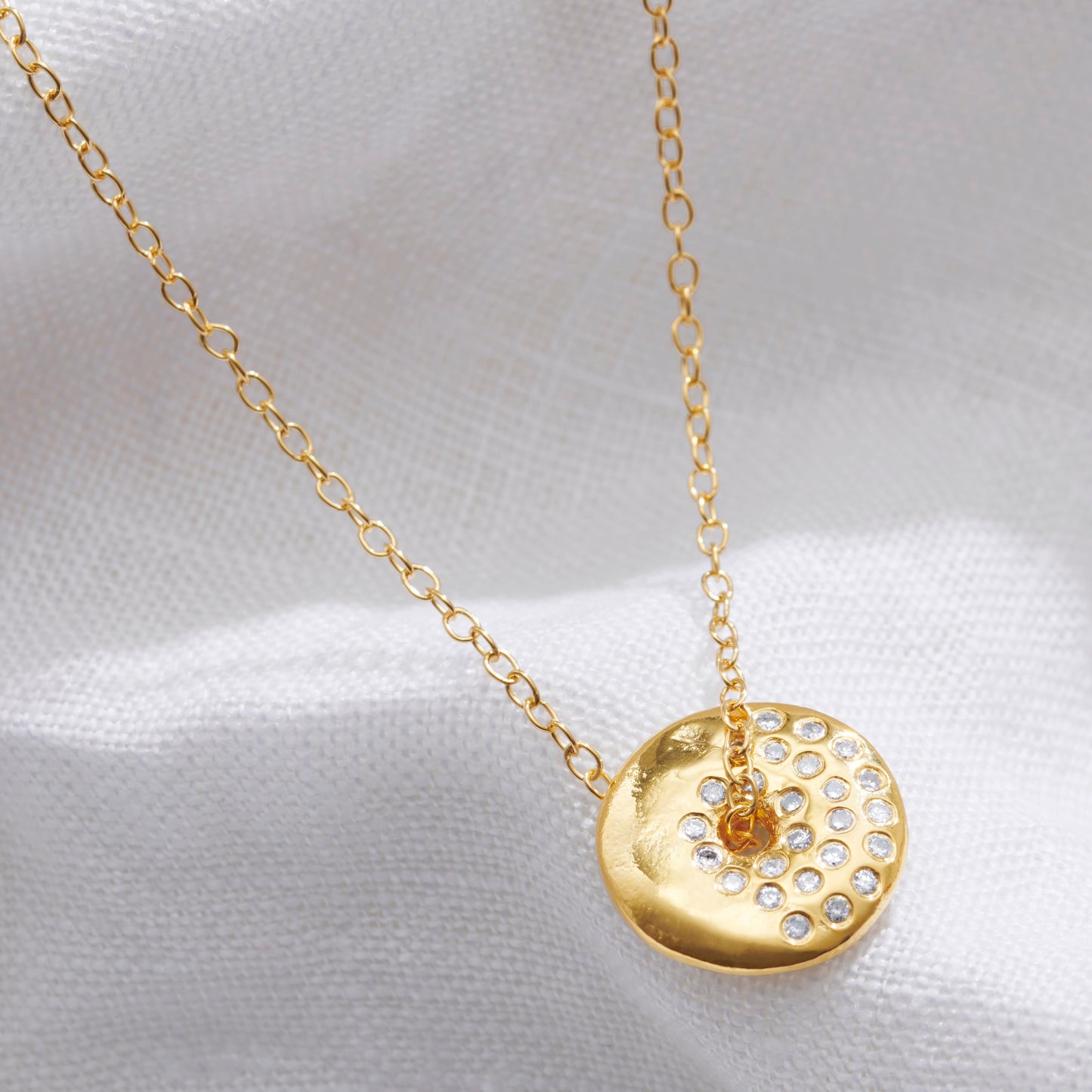 Gold necklace with a round pendant witha textured surface set with white diamonds in the pattern of a waxing gibbous moon on a white fabric background