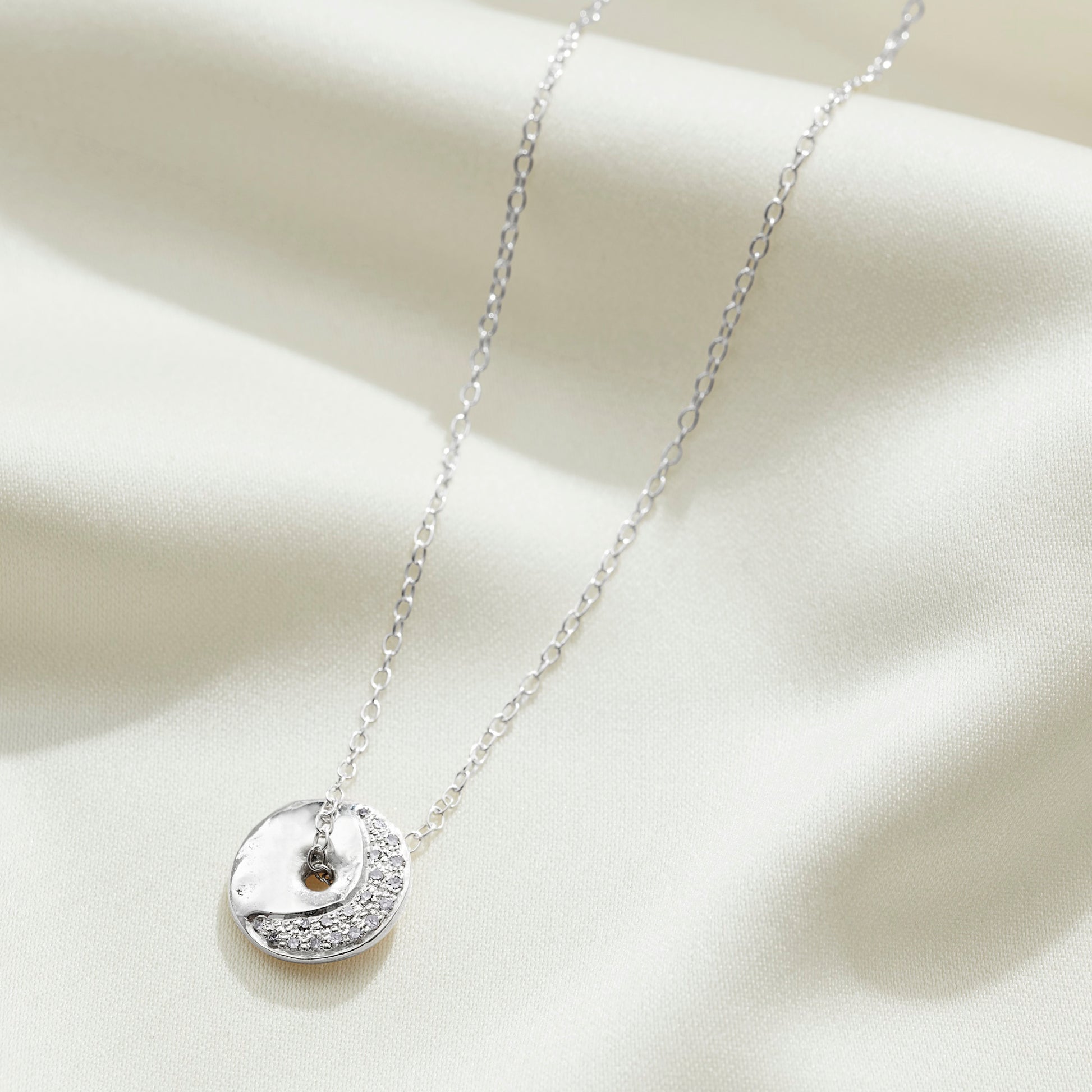 Silver moon phase necklace with a circular pendant witha crescent moon in real diamonds on a light fabric background.