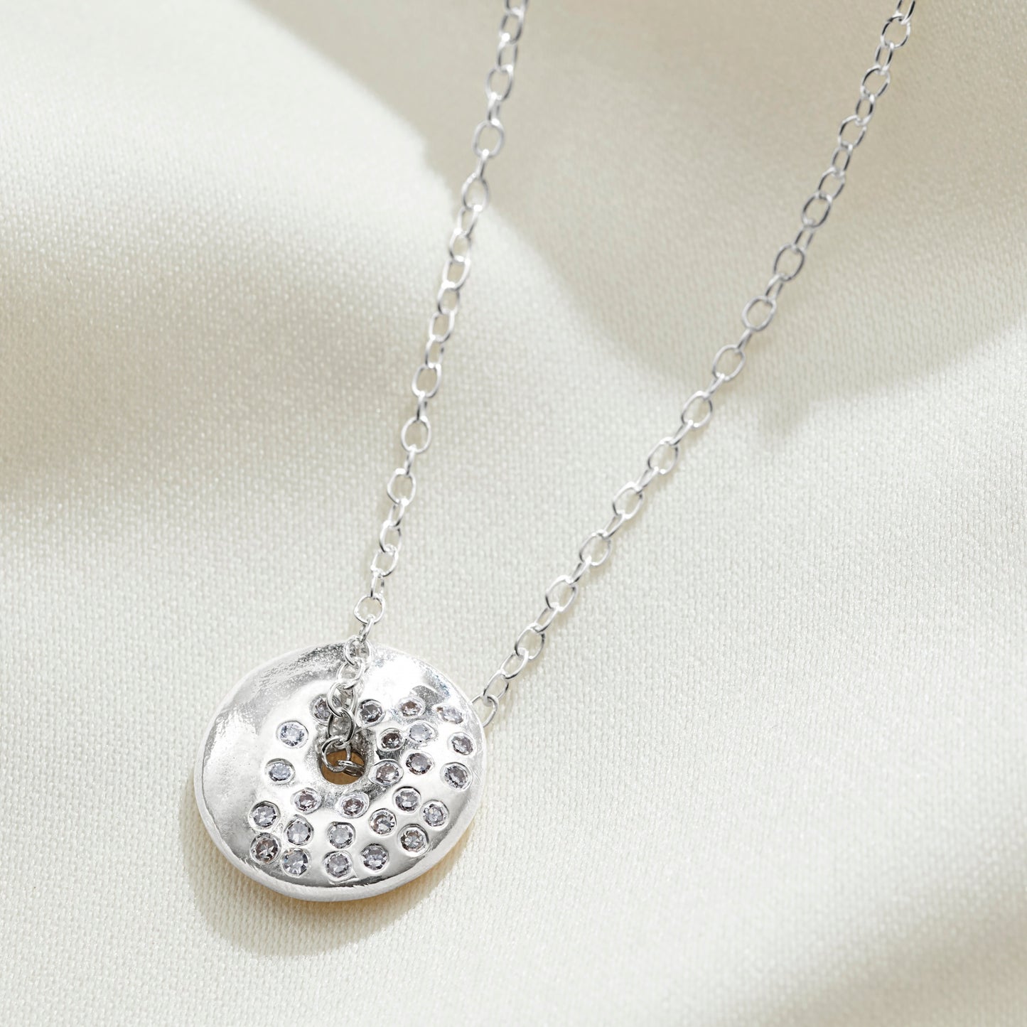 Silver necklace with a round pendant  with a hole in the middle on a light background.  The pendant is set with diamonds to depict the moon phase waxing gibbous.