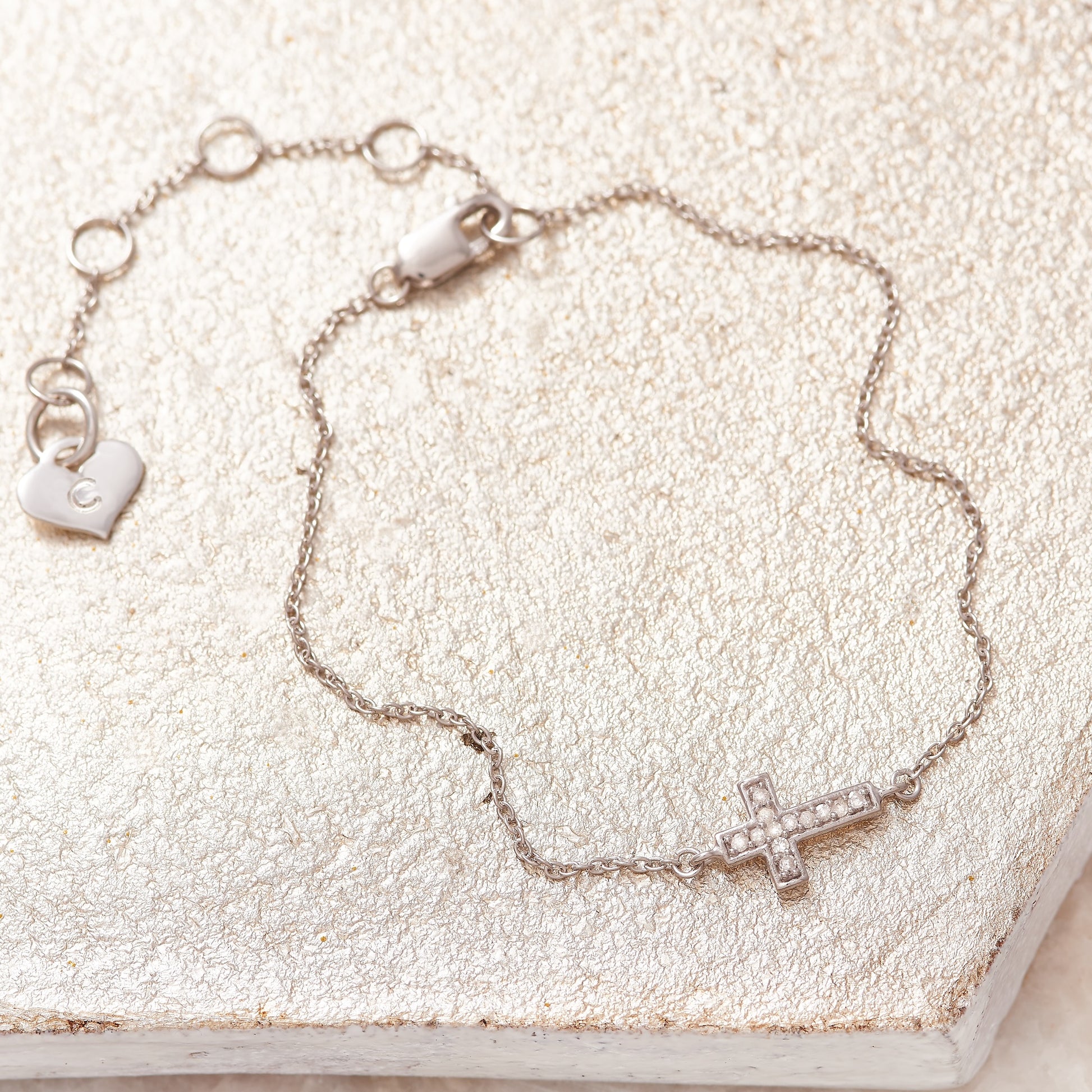 Dainty bracelet with a little cross charm that has been set with tiny white diamonds.  The bracelet is silver with a fine trace chain and fastens with a lobster claw fastening.  The bracelet has an adjustable chain and has a sweet little silver heart charm on the end that has been personalised with the letter C.  Made by Claudette Worters.